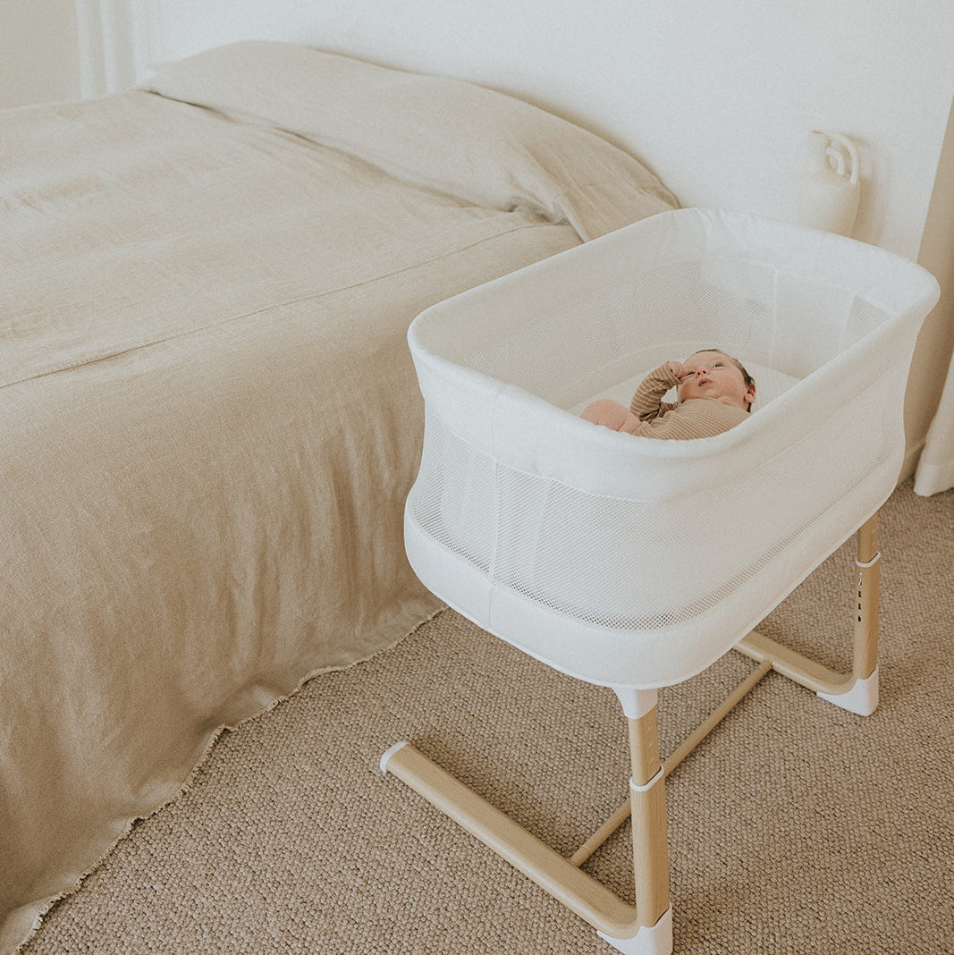 Baby in a white bassinet next to a bed in a bedroom setting