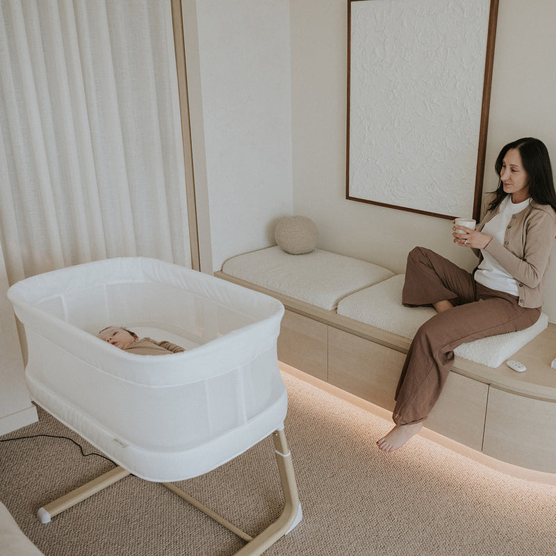 Woman sitting on a bench next to a baby in a bassinet in a room with neutral decor.