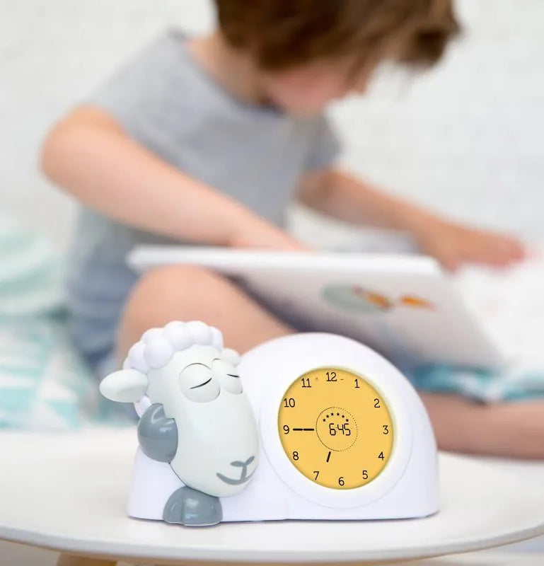 Children's alarm clock with sheep design on a table, child reading book in the background